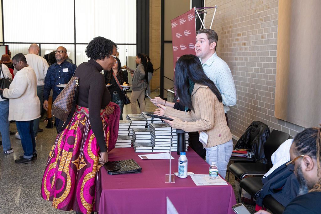 man and woman handing a book to a woman in a brown shirt and pink print skirt