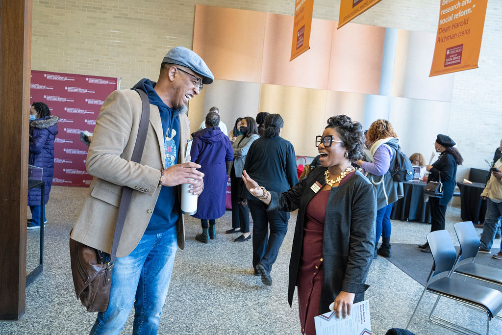 Image of man in brown coat with jeans and grey hat chatting with a woman in a maroon dress and black jacket.
