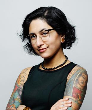 Gabriela Zapata-Alma against a white background wearing glasses, a black top, and showing tattooed arms